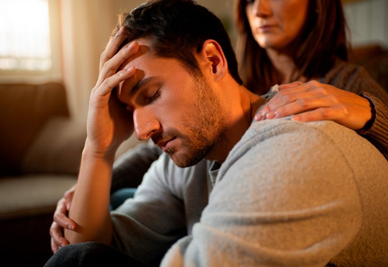 Мужчина в сером свитере сидит, прижимая ладонь ко лбу, за ним стоит женщина, мягко положив руку ему на плечо